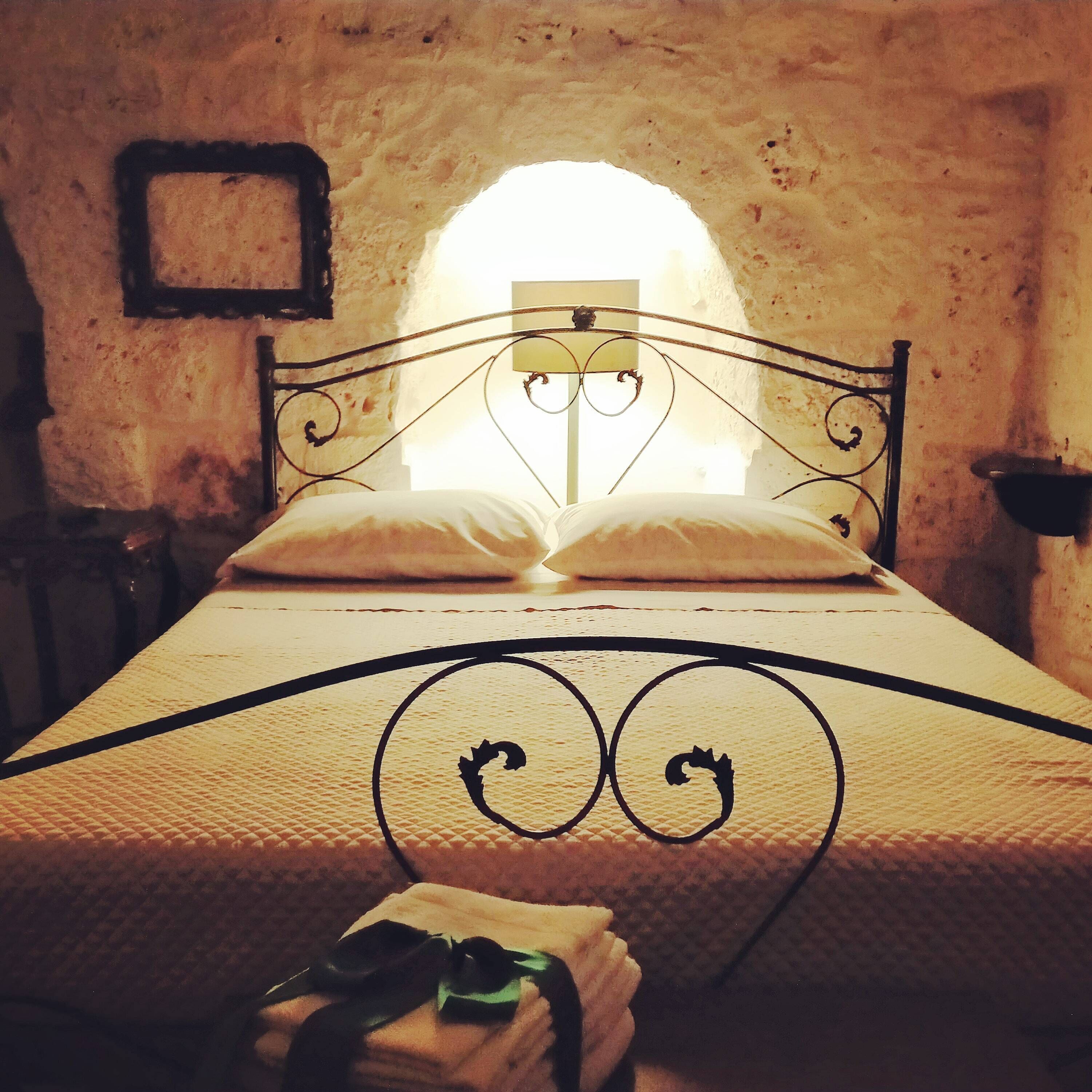 Una cama con estructura metálica y colcha acolchada, una almohada y una mesita de noche con lámpara.