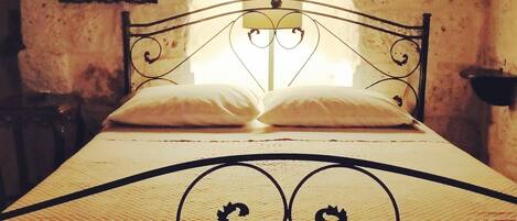 Una cama con estructura metálica y colcha acolchada, una almohada y una mesita de noche con lámpara.