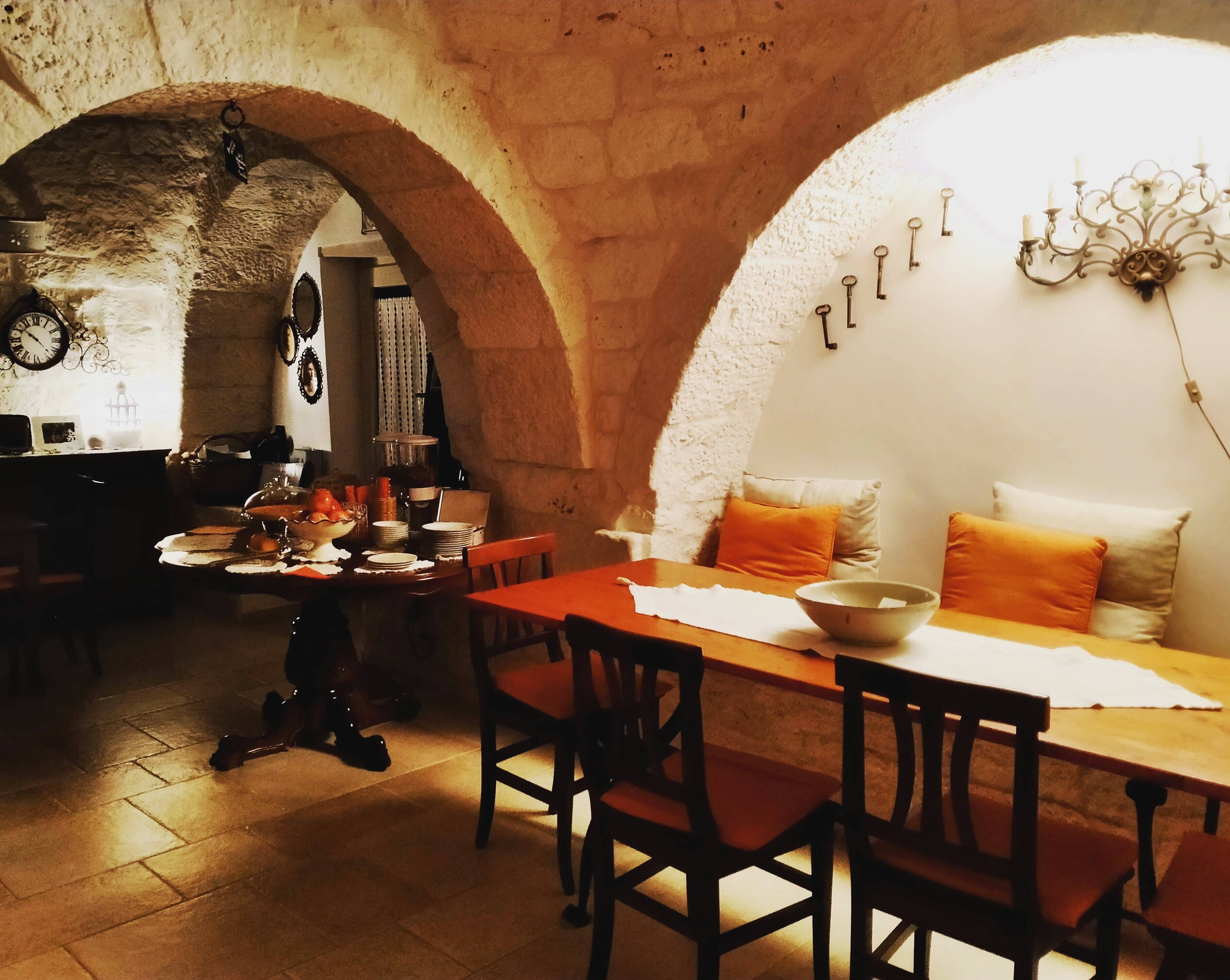 A dining area with a wooden table, chairs, and a chandelier. There is a stone archway and a clock on the wall.