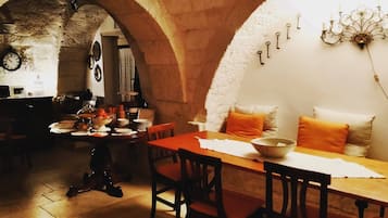 A dining area with a wooden table, chairs, and a chandelier. There is a stone archway and a clock on the wall.