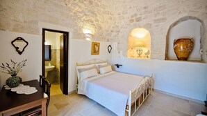 Un dormitorio con techo de piedra, una cama con ropa blanca, una mesita de noche con un florero y una lámpara fijada a la pared.