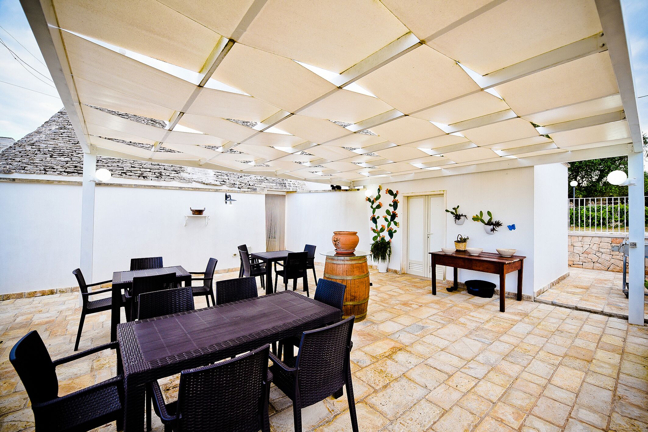 An outdoor patio with a tiled floor, covered by a white pergola, featuring tables and chairs, a wooden barrel, and potted plants.