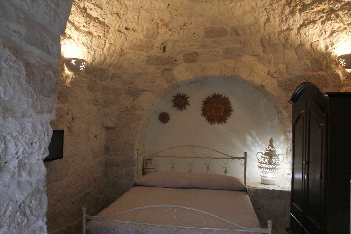 Una habitación con paredes de piedra, una cama, una lámpara de pared y un armario.