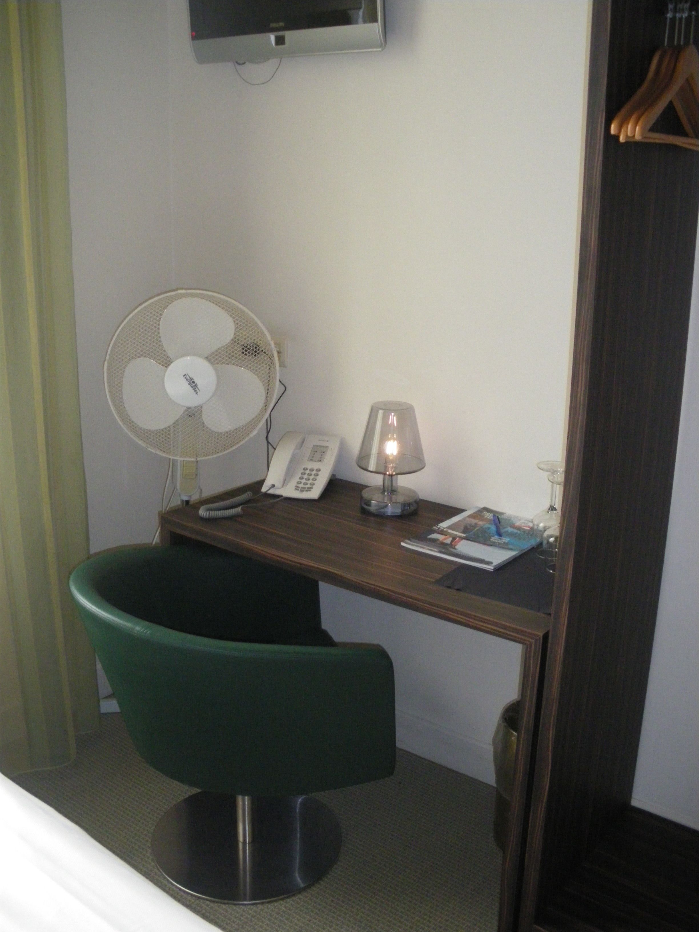 Une chambre d’hôtel avec un bureau en bois, une chaise verte, un ventilateur sur pied, un téléphone, une lampe et un téléviseur fixé au mur.