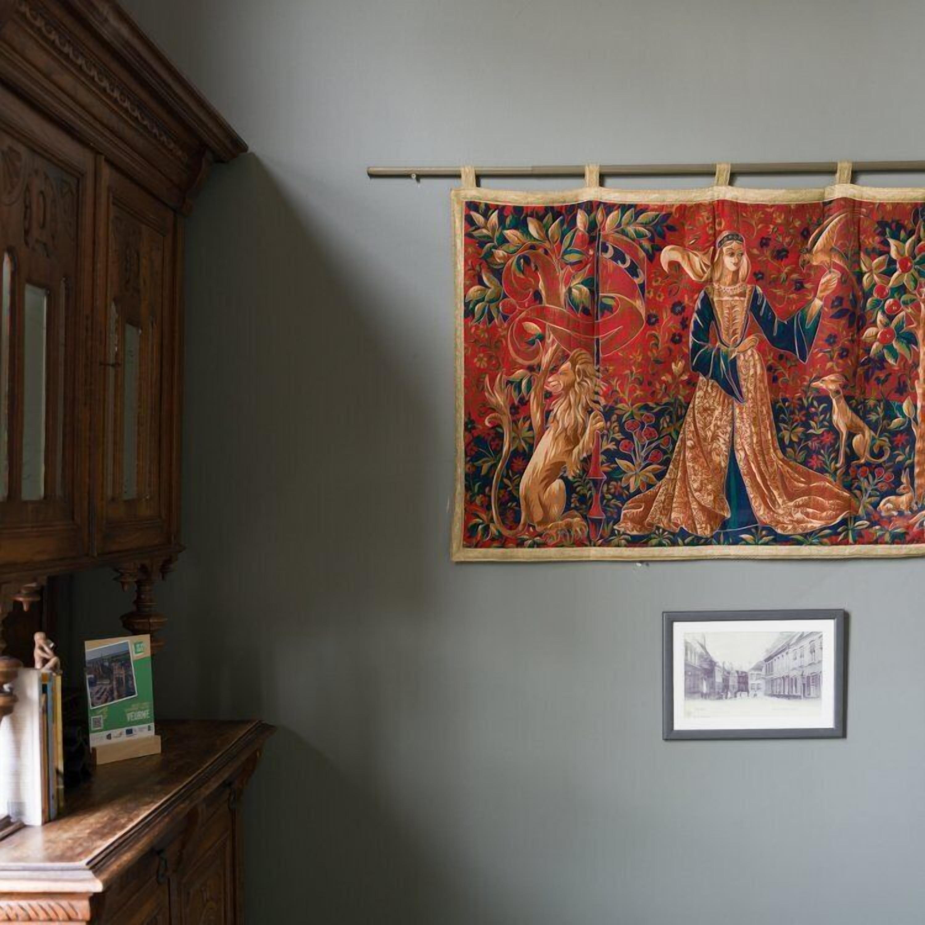 Une tapisserie représentant une figure centrale et un chien, un tableau encadré en dessous, et un meuble en bois sur la gauche.