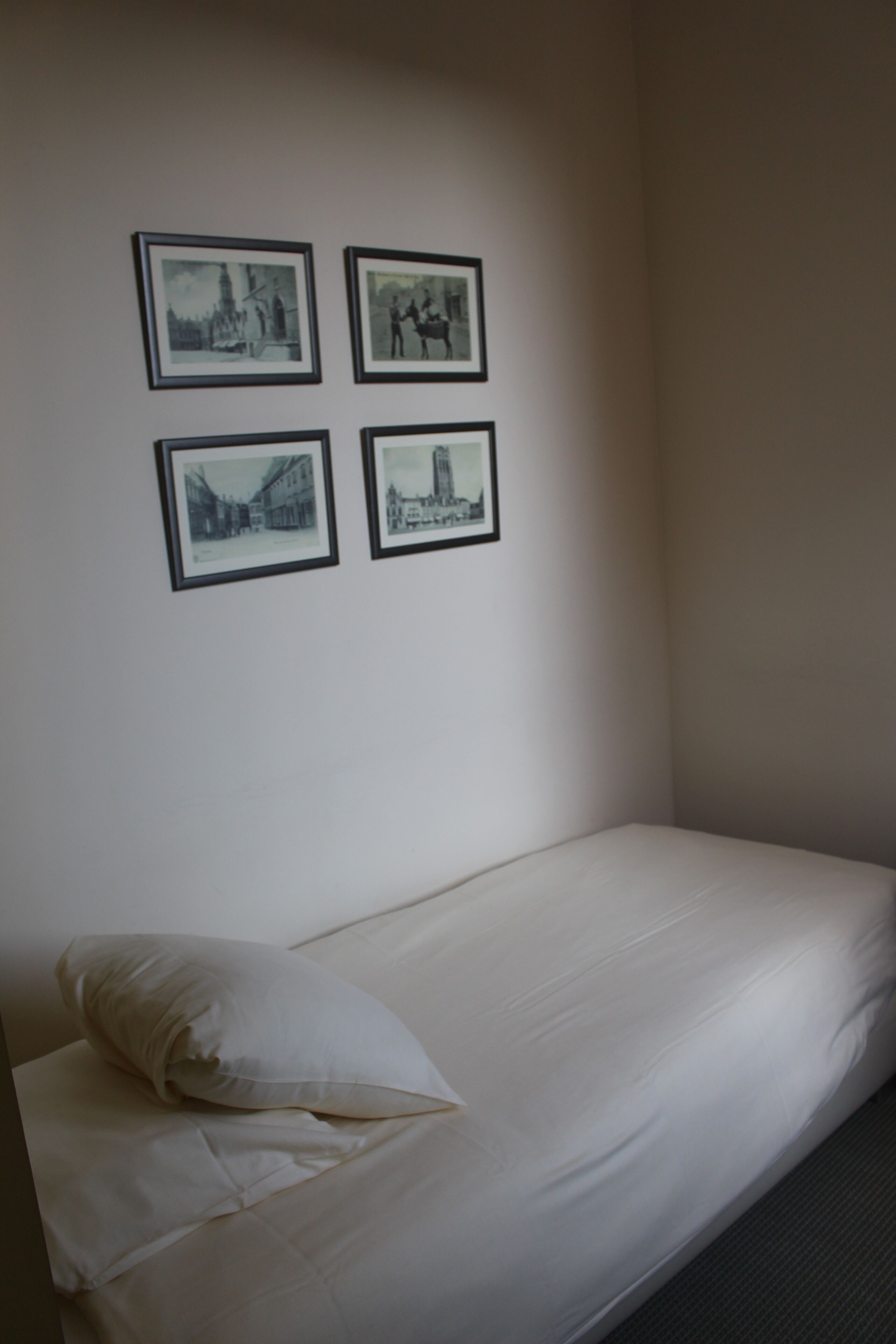 A single bed with white bedding and a pillow, a plain wall with four black-and-white framed pictures, and a plain wall.