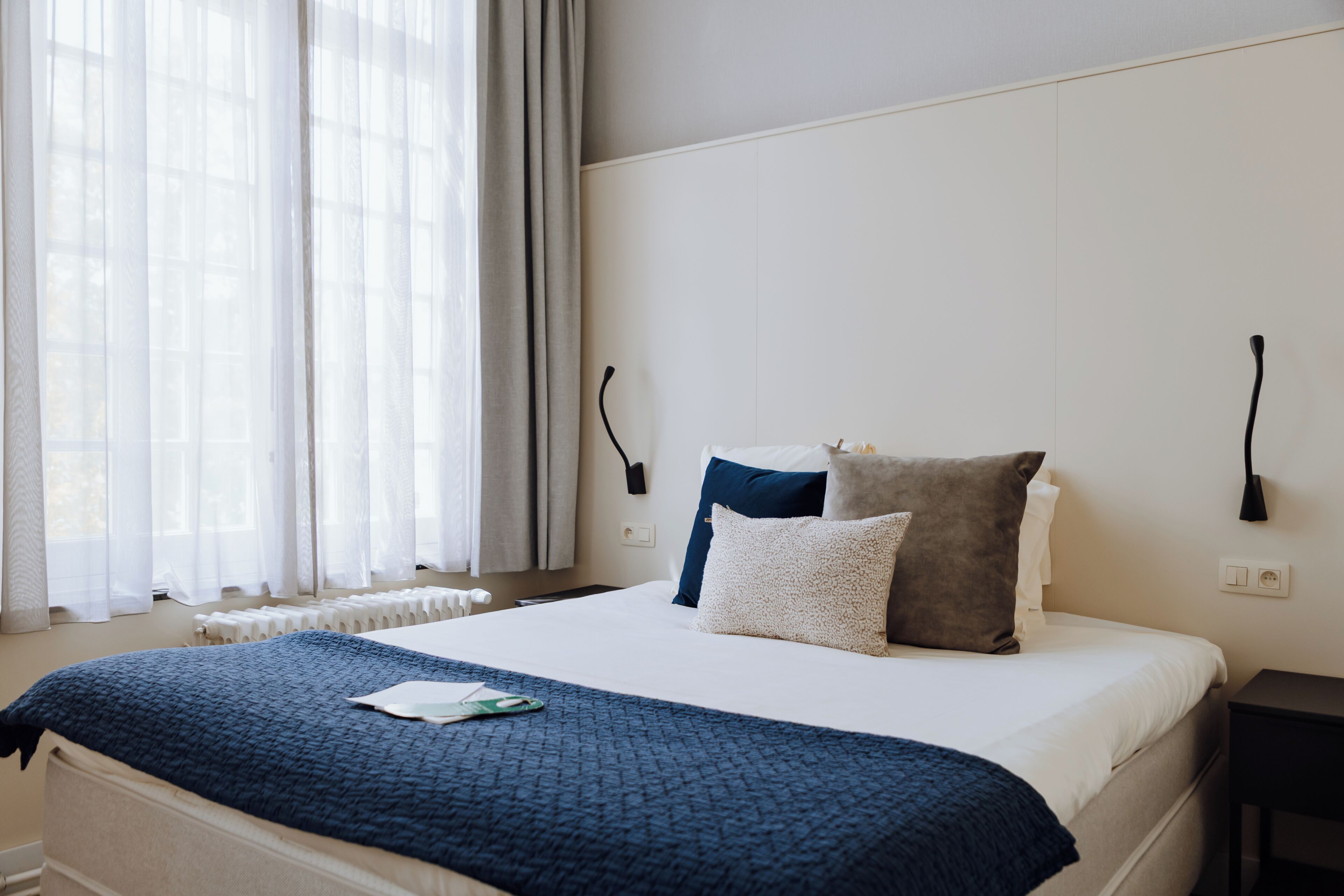 A neatly made bed with a blue blanket, a white mattress, and two pillows in shades of blue and gray.