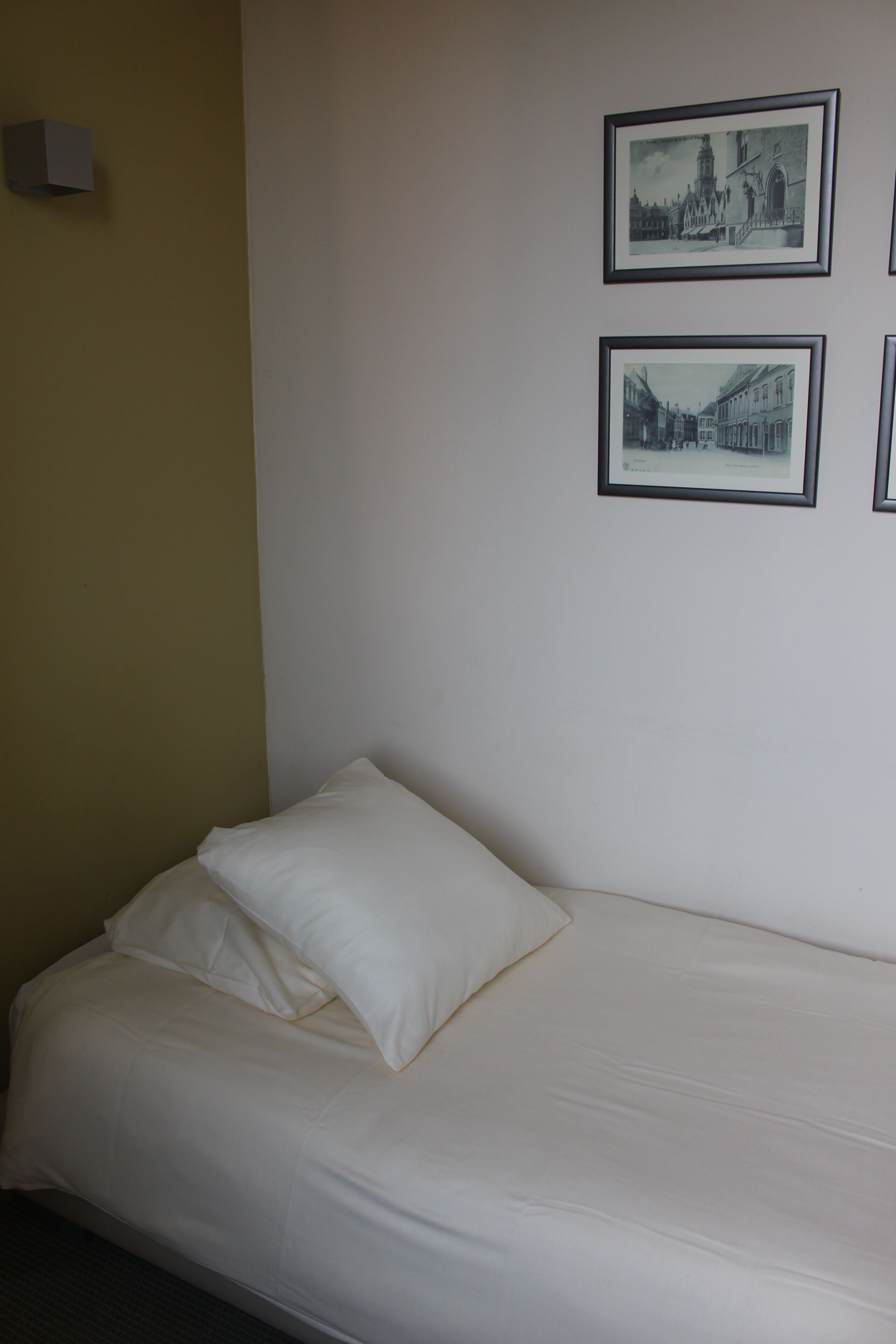 Un lit simple avec une literie blanche dans une pièce aux murs unis. Deux photographies en noir et blanc encadrées sont accrochées au mur.