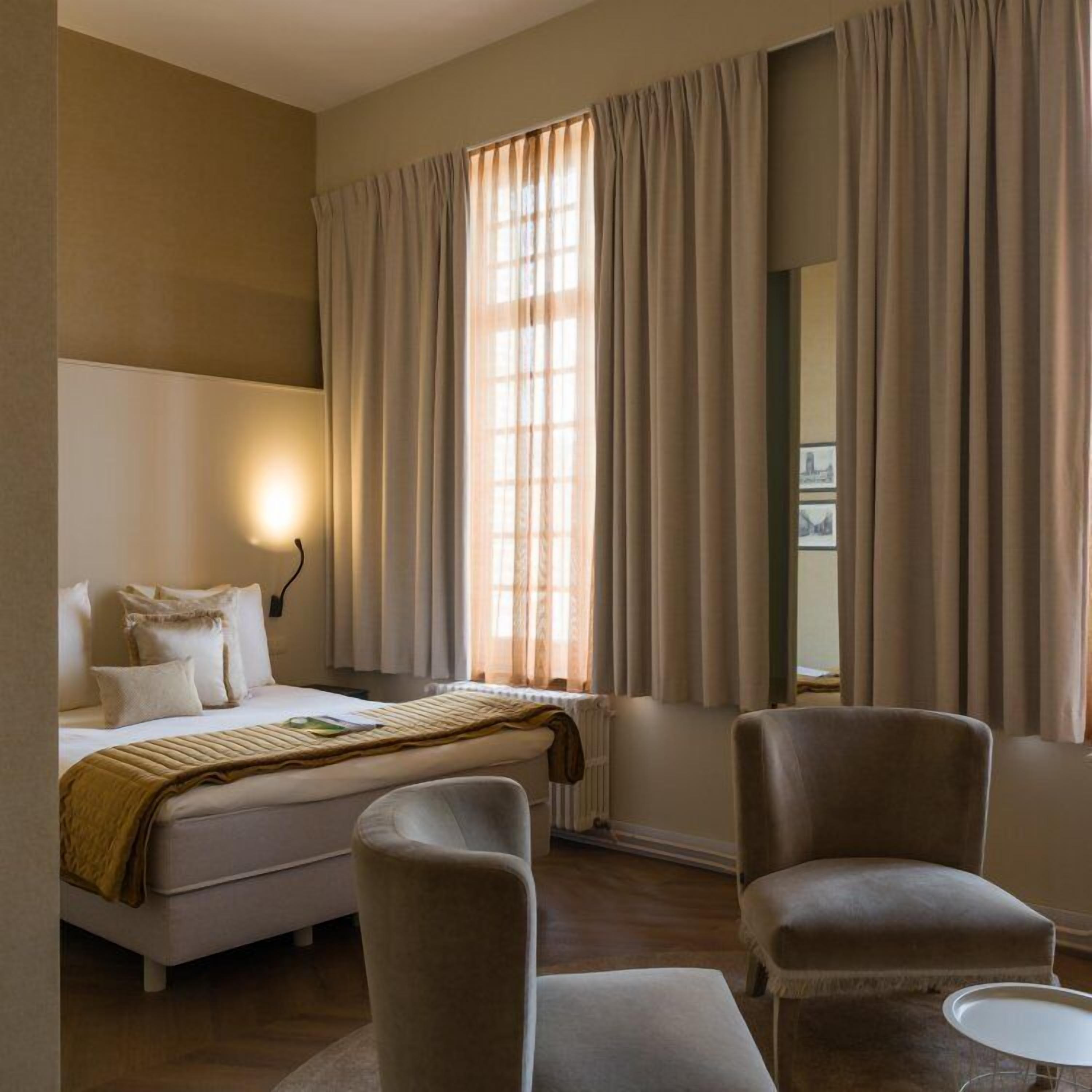 A hotel room with a bed, two armchairs, a small table, and a wall-mounted lamp.