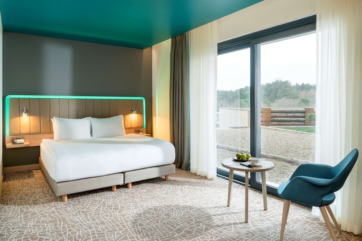 Een moderne hotelkamer met een groot bed, een nachtkastje met een lamp, een klein rond tafeltje met een schaal fruit, en een blauwe stoel.