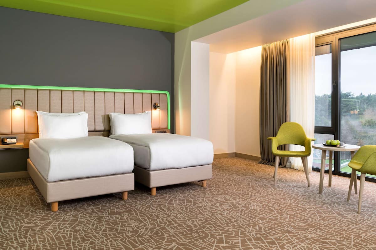 Een hotelkamer met twee bedden, een nachtkastje, een lamp, een stoel, een tafel en uitzicht op het omliggende landschap.