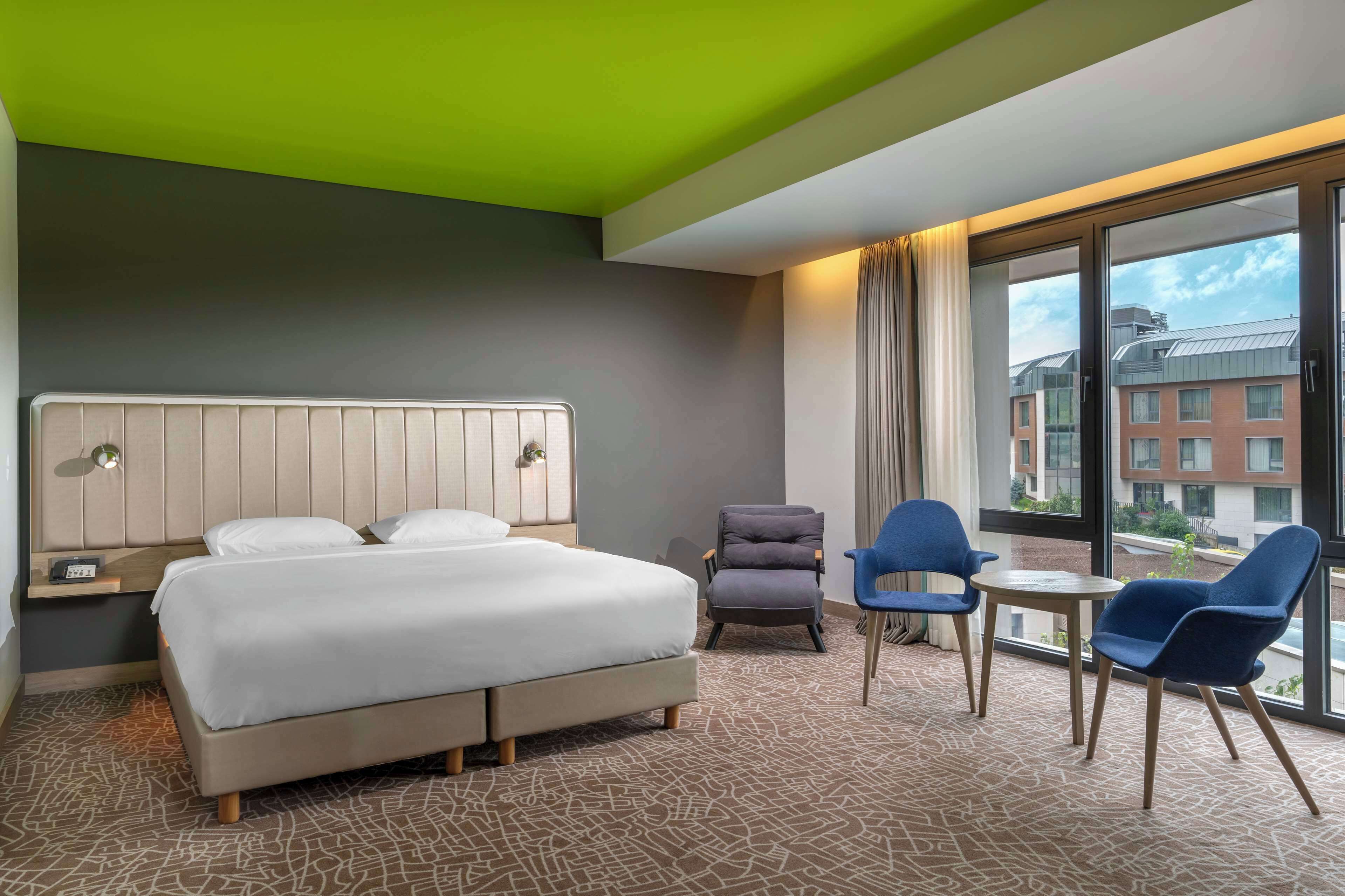 A hotel room with a bed, a chair, a table, and a view of buildings through large windows.