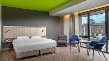 A hotel room with a bed, a chair, a table, and a view of buildings through large windows.