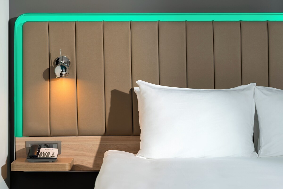 A hotel room with a padded headboard, a bedside table with a phone, and white bedding.