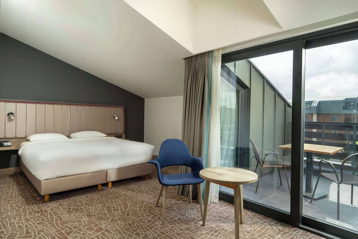 Een hotelkamer met een groot bed, een blauwe stoel, een klein tafeltje en een balkon met uitzicht op gebouwen.