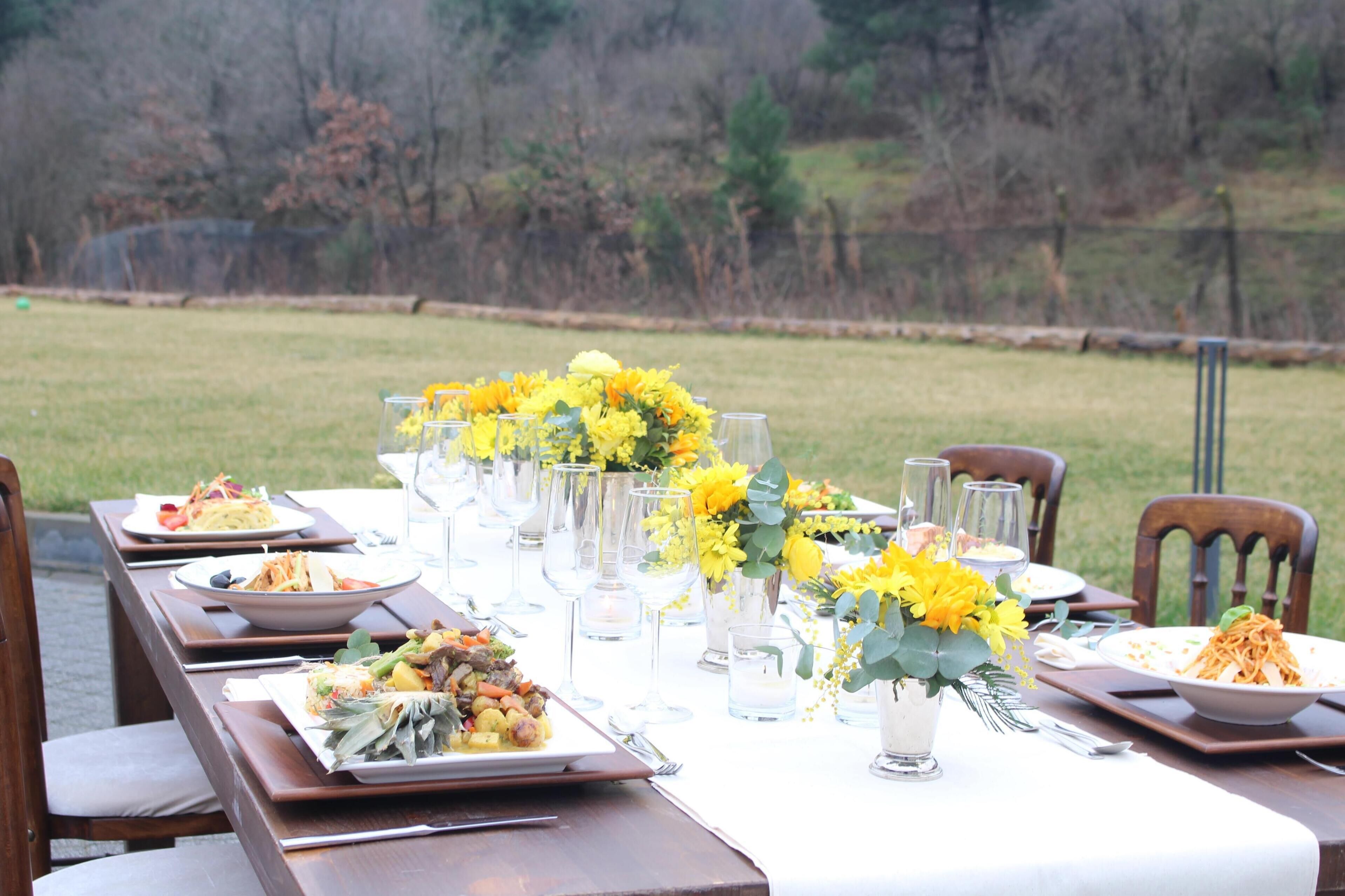 A dining table set outdoors with multiple plates of food, glasses, and yellow flower arrangements.