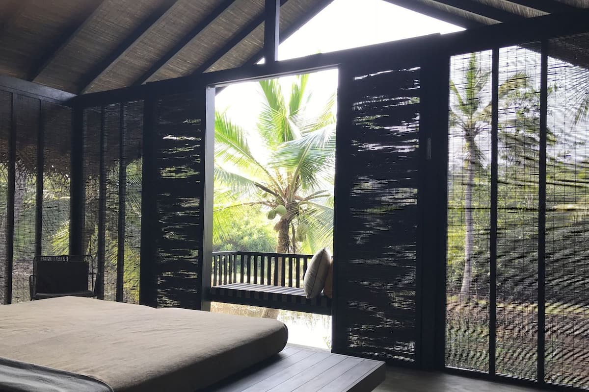 A room with a bed, a bench, and a view of palm trees through a window.