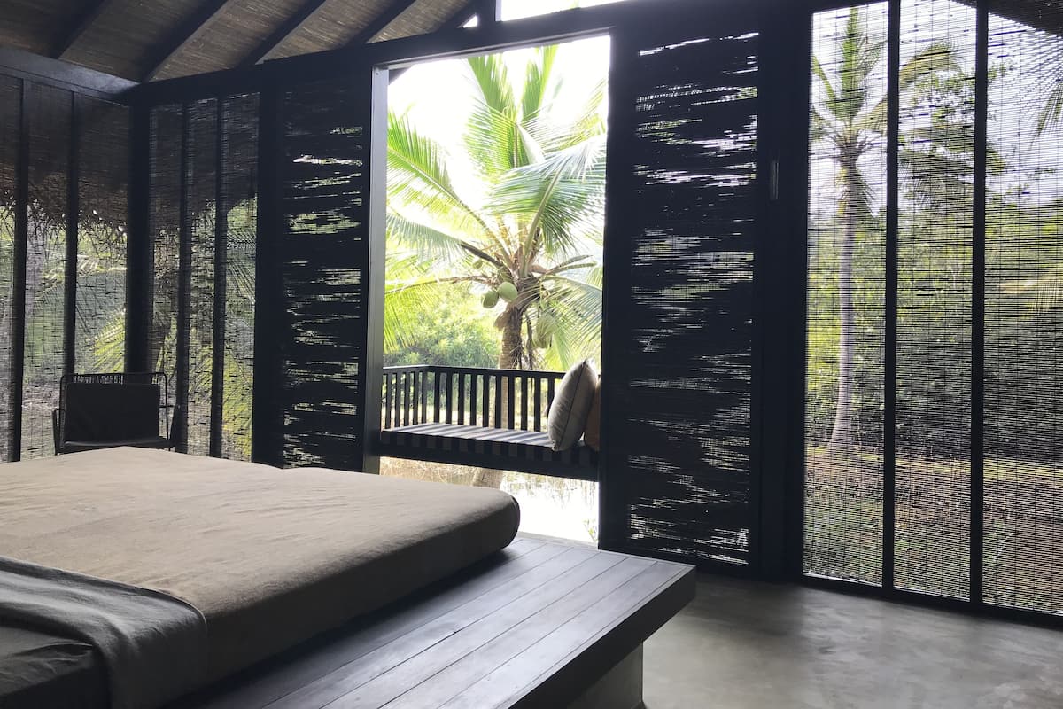 A room with a bed, a bench, and a view of palm trees through a window with a black mesh screen.