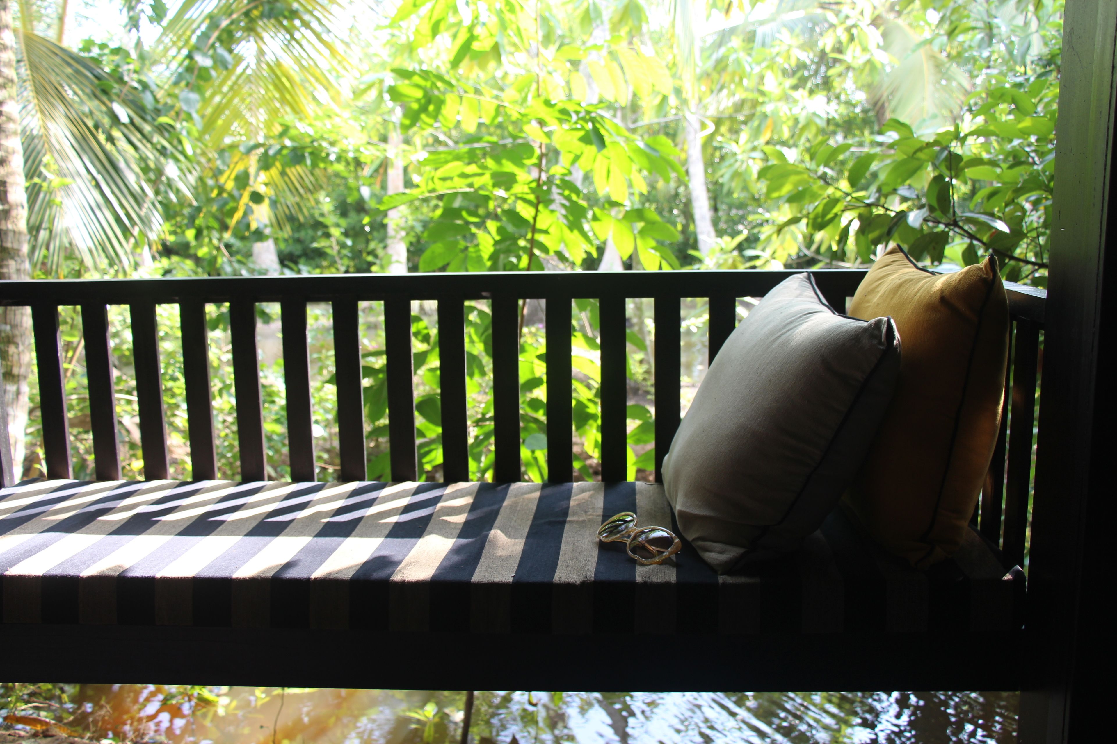 A wooden bench with striped cushions and a cushion with a tassel, positioned by a window with a view of lush greenery.