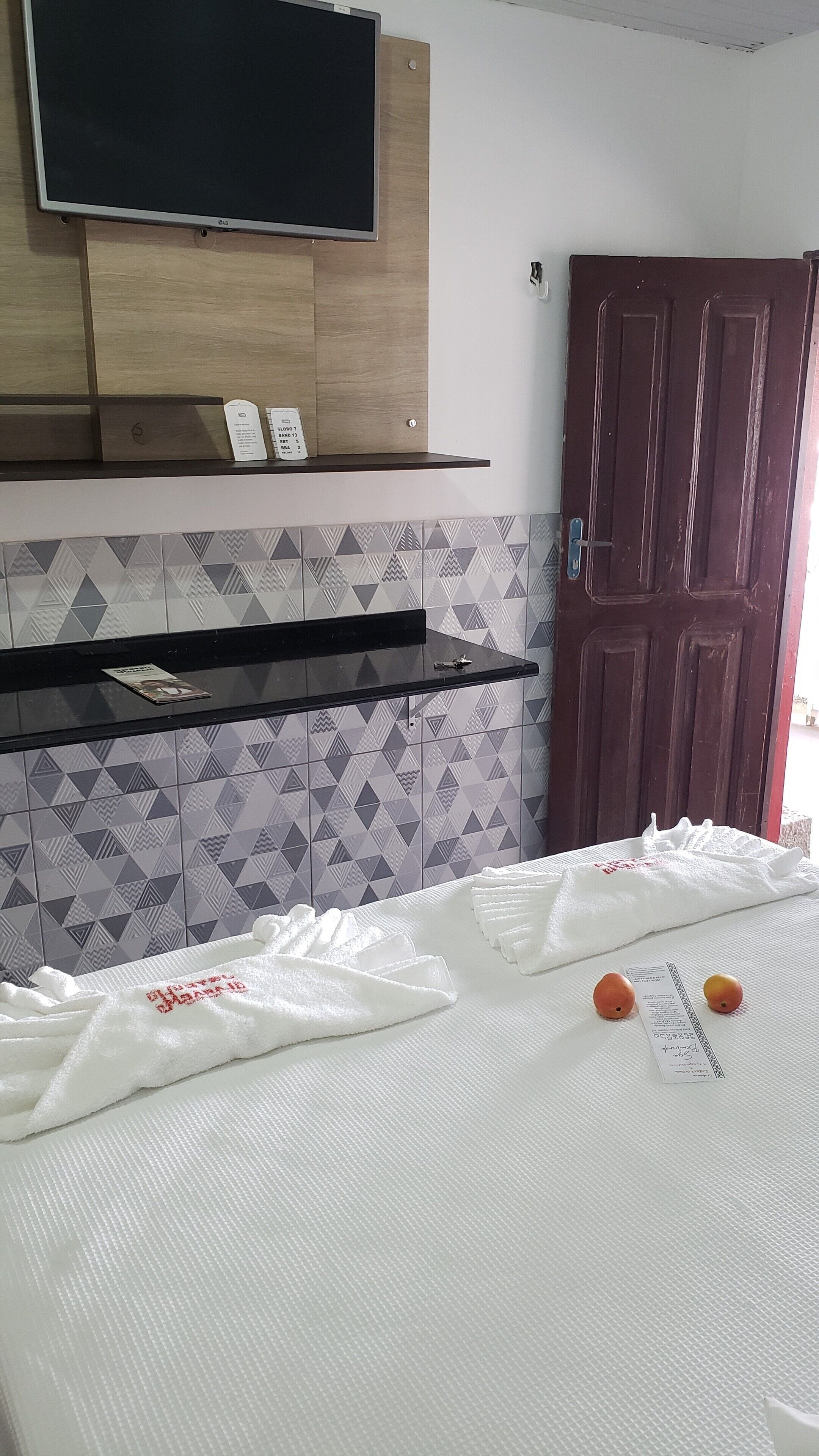 A hotel room with a bed, a TV mounted on a wooden panel, a shelf with a remote control, and a door.