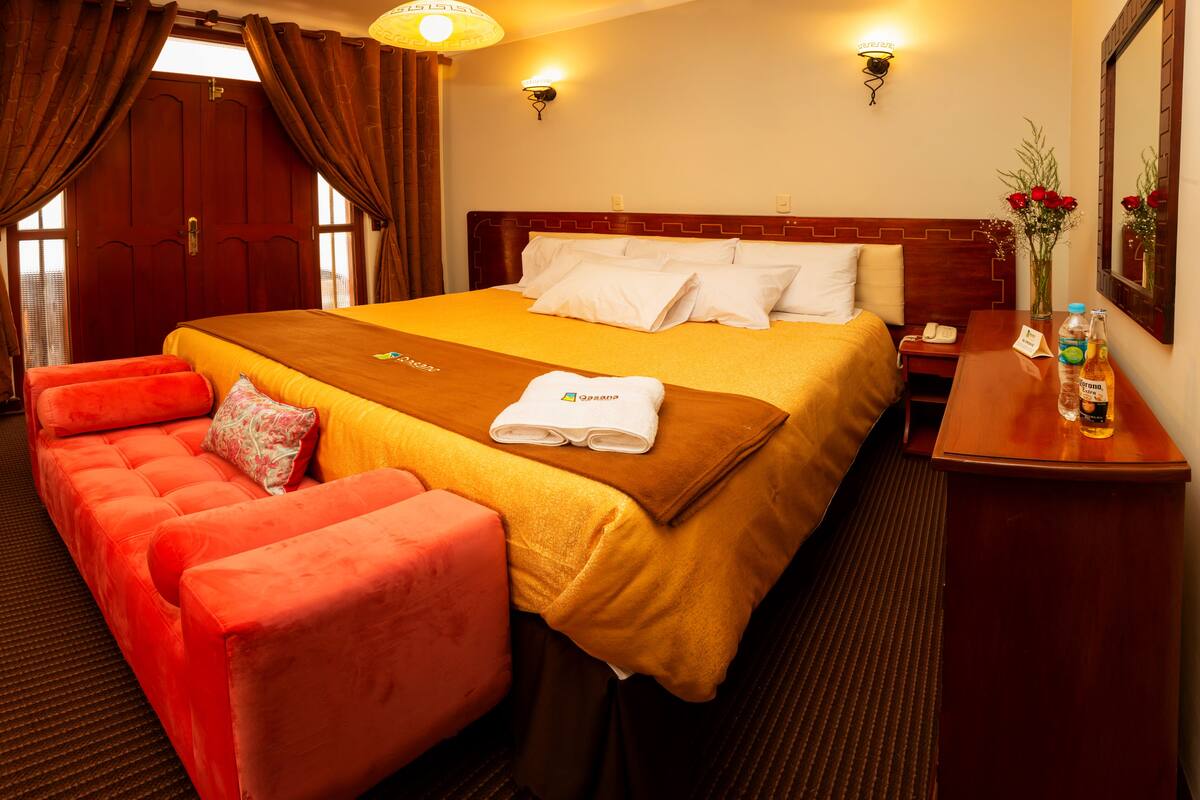 Une chambre d’hôtel avec un grand lit, une tête de lit en bois, un canapé rouge, une table de chevet en bois et un vase rempli de fleurs.