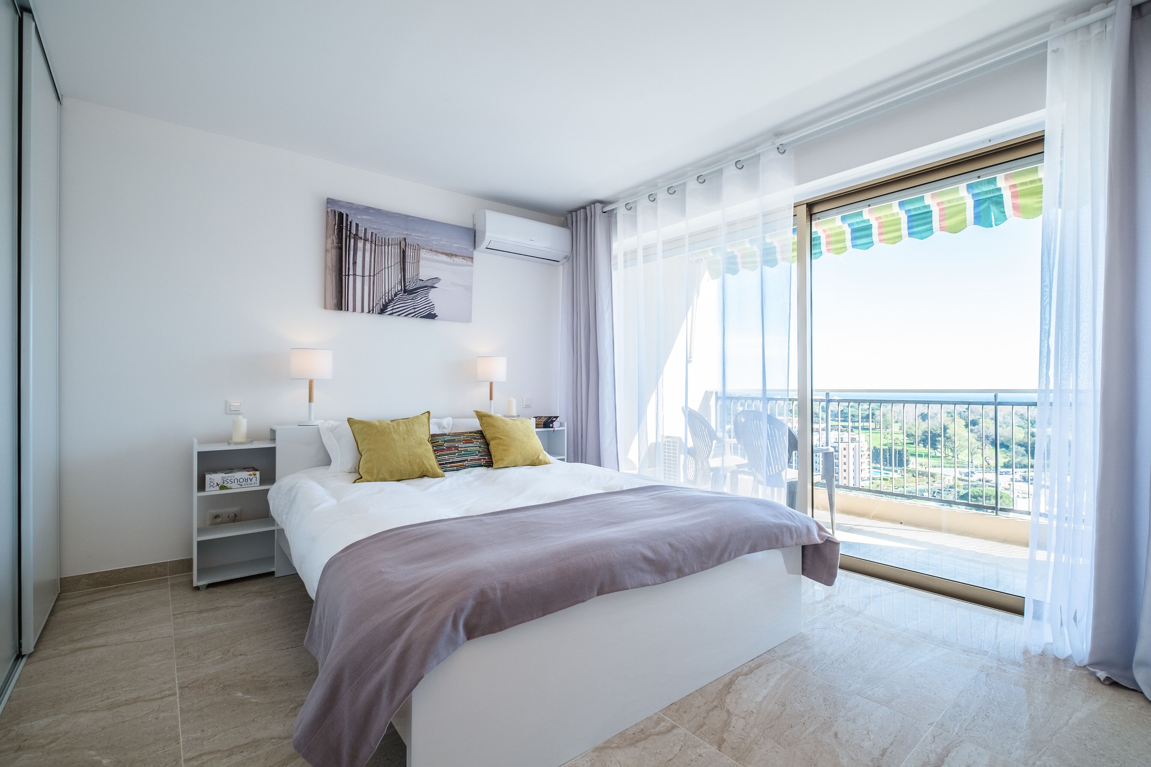 A modern hotel room with a large bed, a nightstand, a wall-mounted air conditioner, and a balcony with a view of the city.