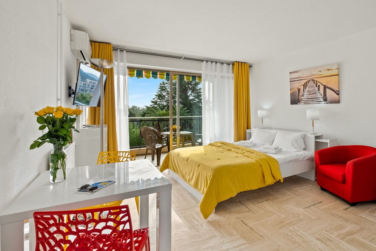 Habitación de hotel con una cama, una silla roja, una mesa con un florero y un refrigerador, además de un televisor.