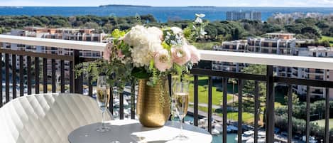 A balcony with a table set for two, a vase of flowers, and a view of a marina and buildings.