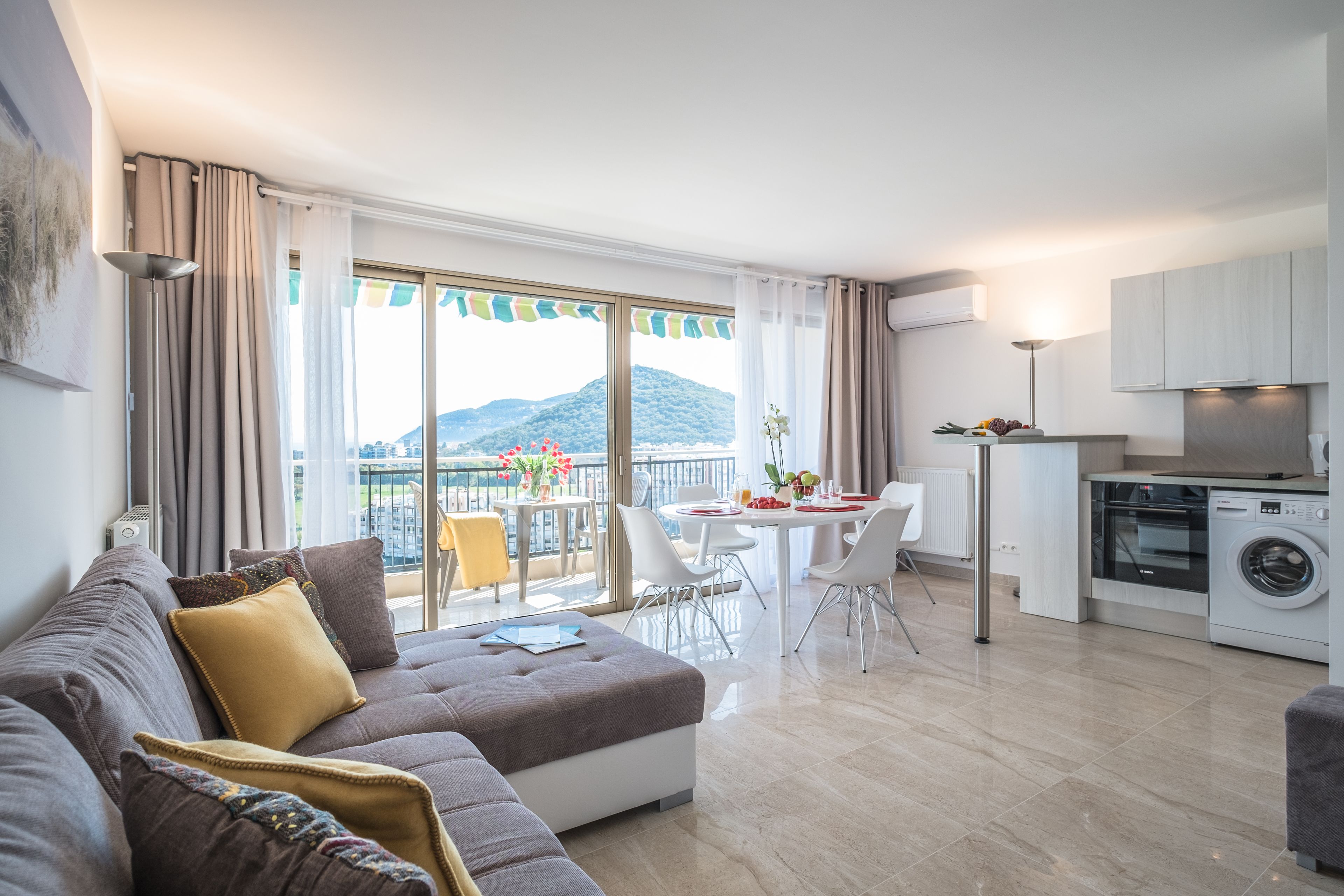 Un appartement moderne comprenant un salon, un coin repas et une kitchenette. Il dispose d’une grande fenêtre offrant une vue sur les montagnes et d’un balcon.