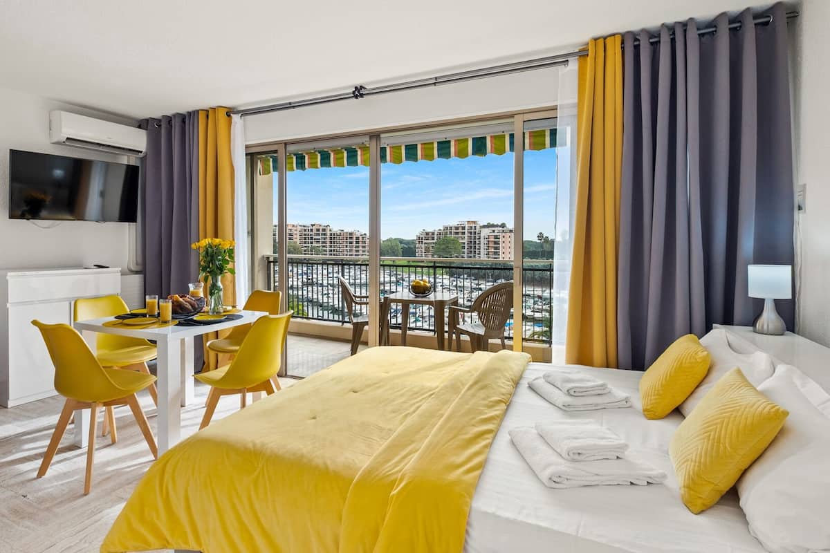 Habitación de hotel con cama, escritorio con sillas, televisor y balcón con vistas a edificios.