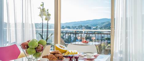 Una mesa de desayuno con frutas, pasteles y bebidas, con vistas a un paisaje pintoresco.