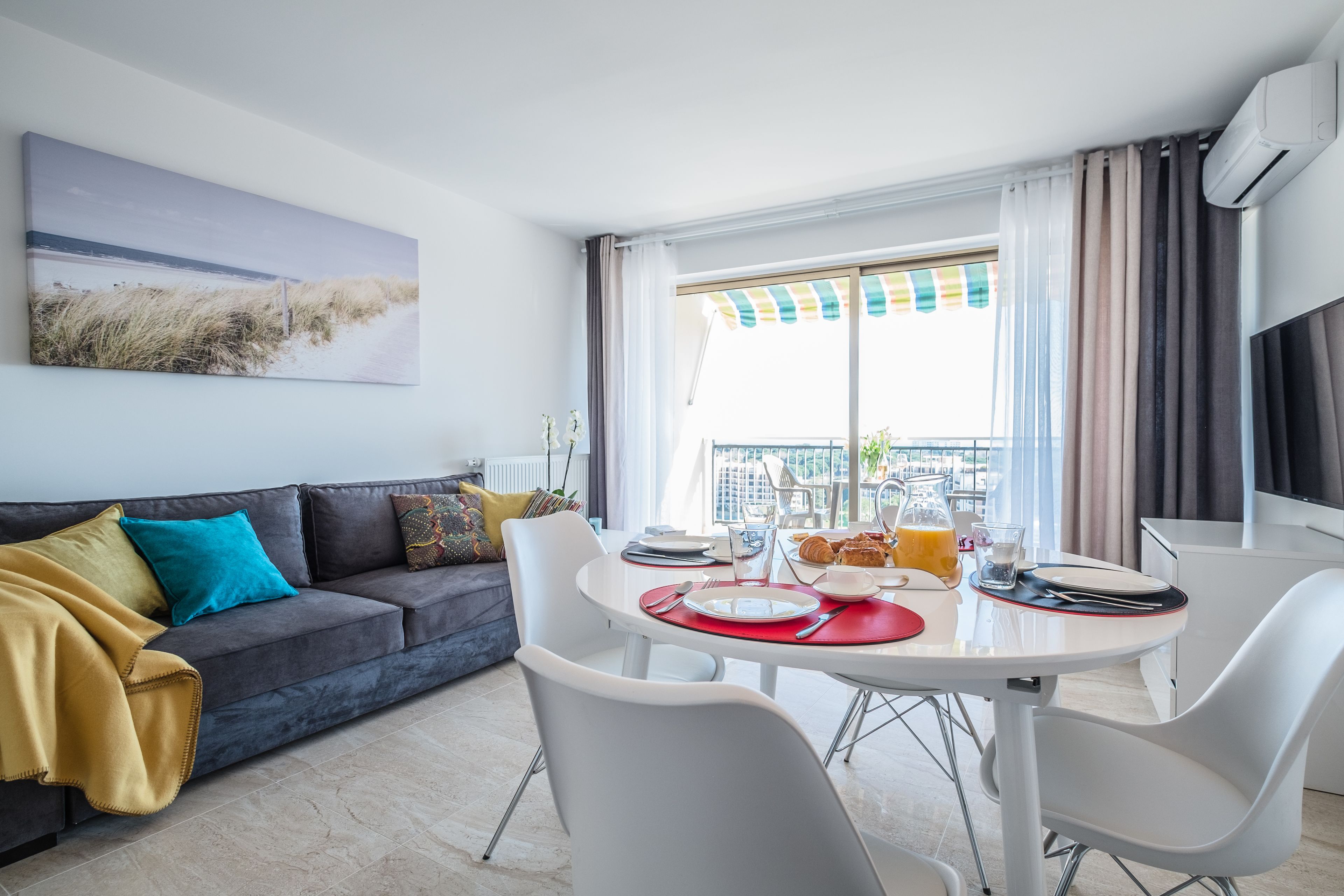Een moderne woonkamer met een grijze bank, een eettafel met stoelen en een groot raam met gordijnen.