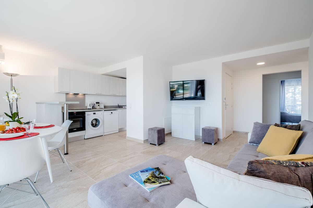 Una sala de estar moderna con un sofá gris, una mesa de comedor con individuales rojos, una cocina al fondo y un televisor montado en la pared.