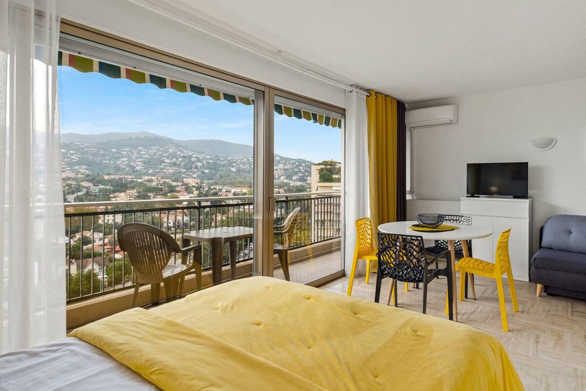 Habitación de hotel con balcón, cama, sofá, televisor y zona de comedor con sillas y mesa.