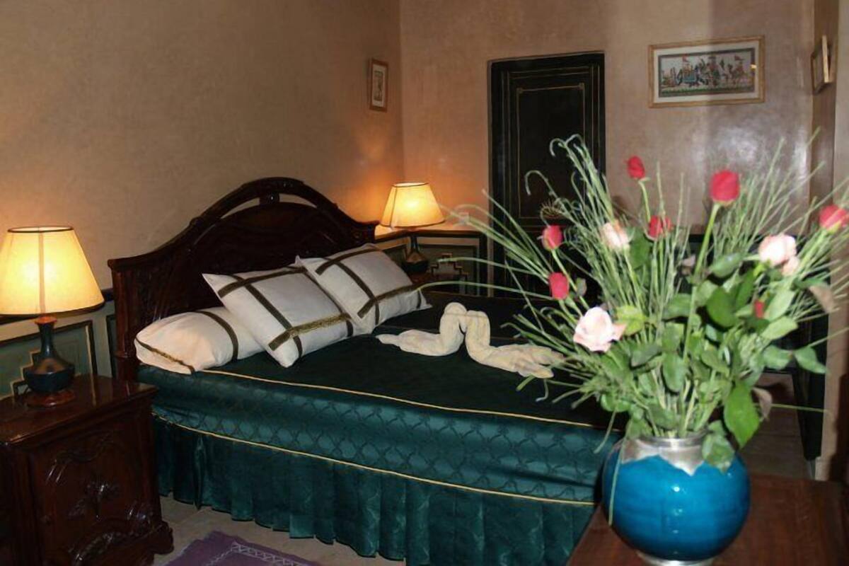 A neatly made bed with a dark green bedspread and white pillows, a vase with flowers on a bedside table, and a framed picture on the wall.