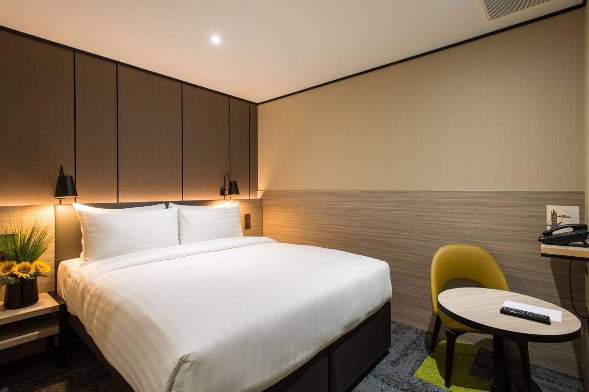 A modern hotel room with a neatly made bed, a small desk with a chair, and a wall-mounted phone.