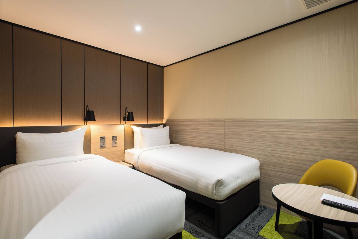 A hotel room with two beds, a small table, a chair, and a wall-mounted lamp.