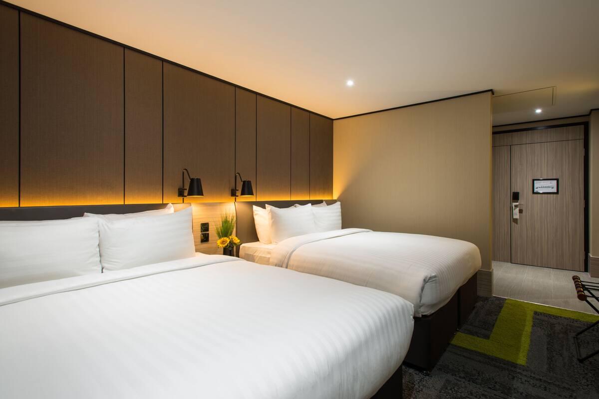 A hotel room with two beds, a nightstand with lamps, a mirror, and a door with a light switch.