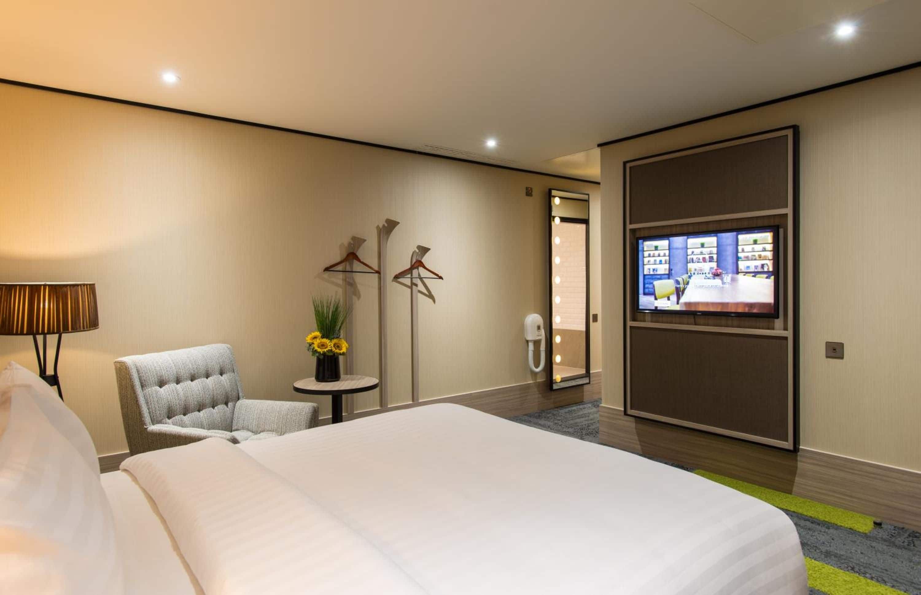 A hotel room with a large bed, a chair, a small table with a vase of flowers, a mirror with lights, and a television on the wall.