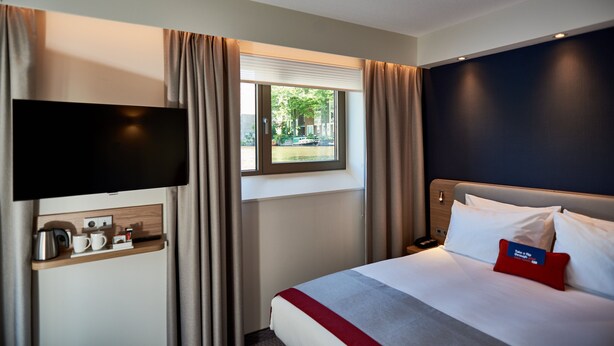 A hotel room with a bed, a TV mounted on the wall, a window with curtains, and a small table with a kettle and cups. - Holiday Inn Express Amsterdam - City Hall by IHG (Amsterdam)
