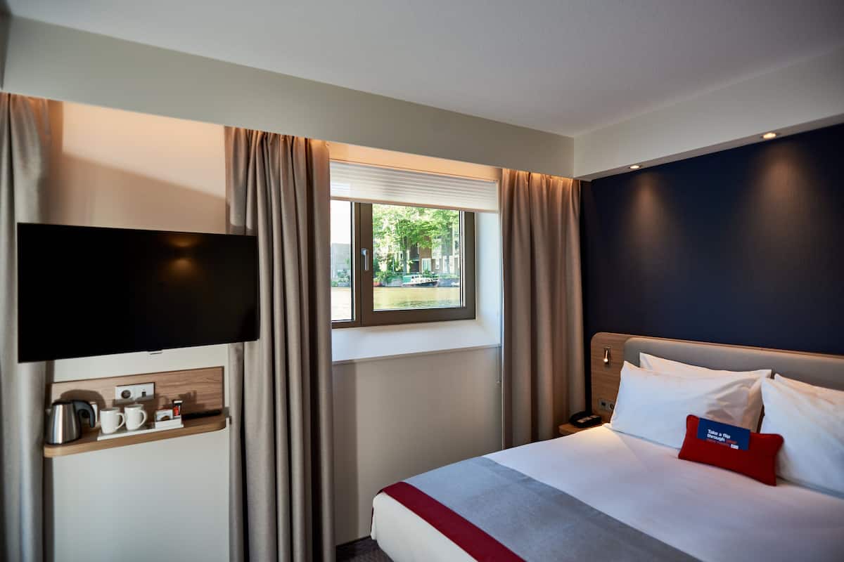 A hotel room with a bed, a TV mounted on the wall, a window with curtains, and a small table with a kettle and cups.