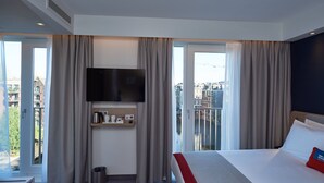 A hotel room with a bed, a TV mounted on the wall, a small shelf with toiletries, and a view of the city through large windows. - Holiday Inn Express Amsterdam - City Hall by IHG (Amsterdam)