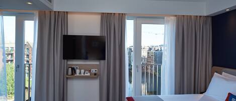 A hotel room with a bed, a TV mounted on the wall, a small shelf with toiletries, and a view of the city through large windows.