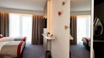 Une chambre d’hôtel avec deux lits, un bureau avec un miroir, une télévision et une poubelle.