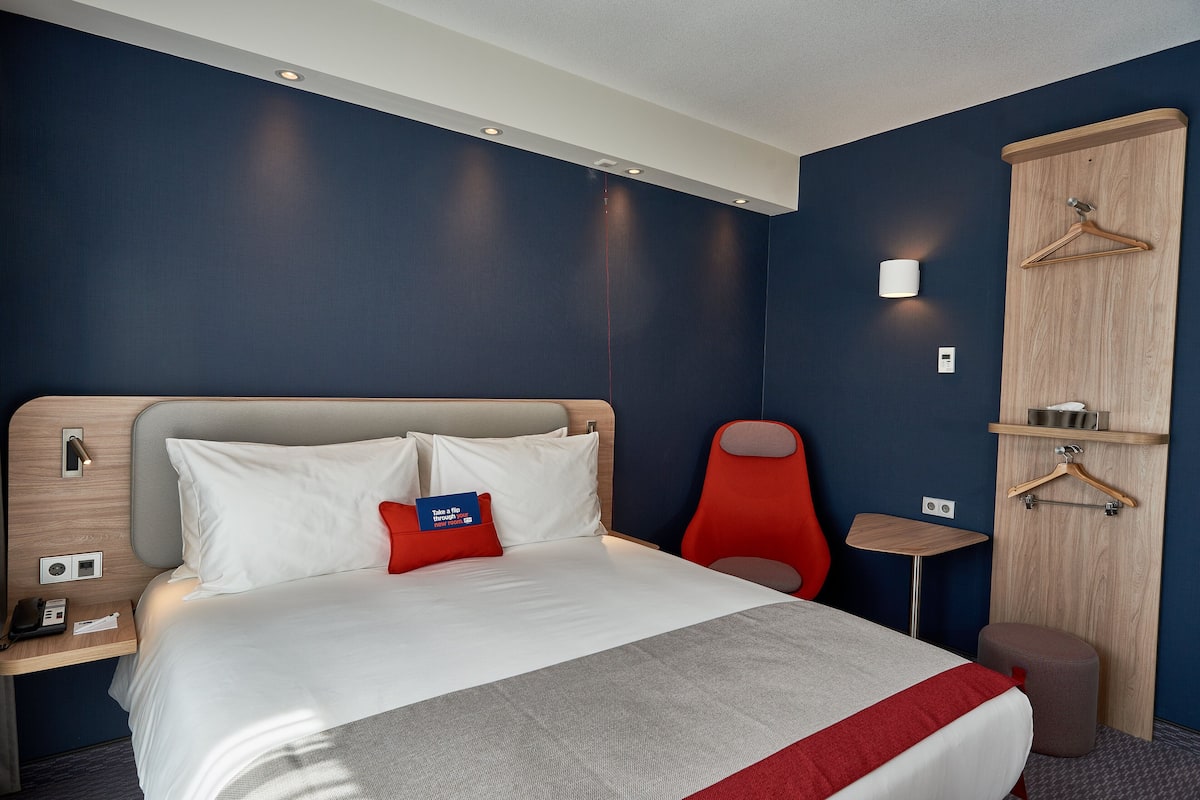A hotel room with a bed, a red chair, a small table, a wooden wardrobe, and a wall-mounted light.