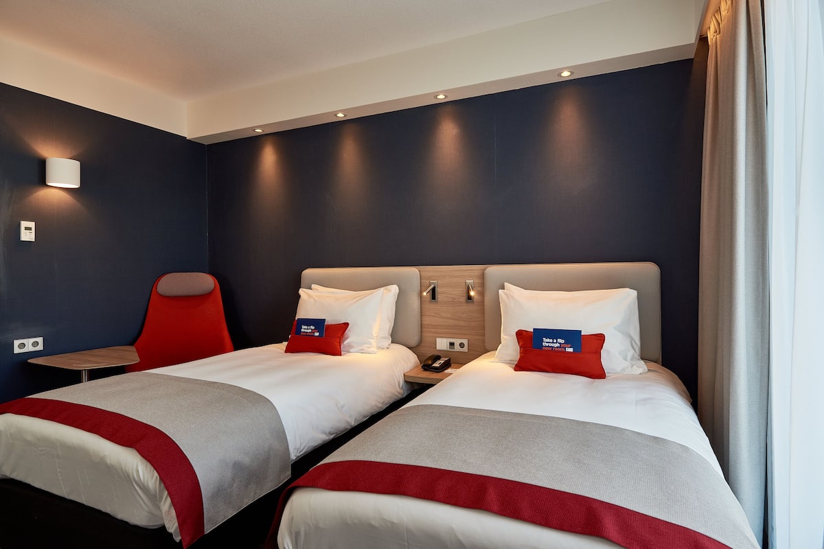 A hotel room with two beds, a red chair, a small table, and a wall-mounted phone.