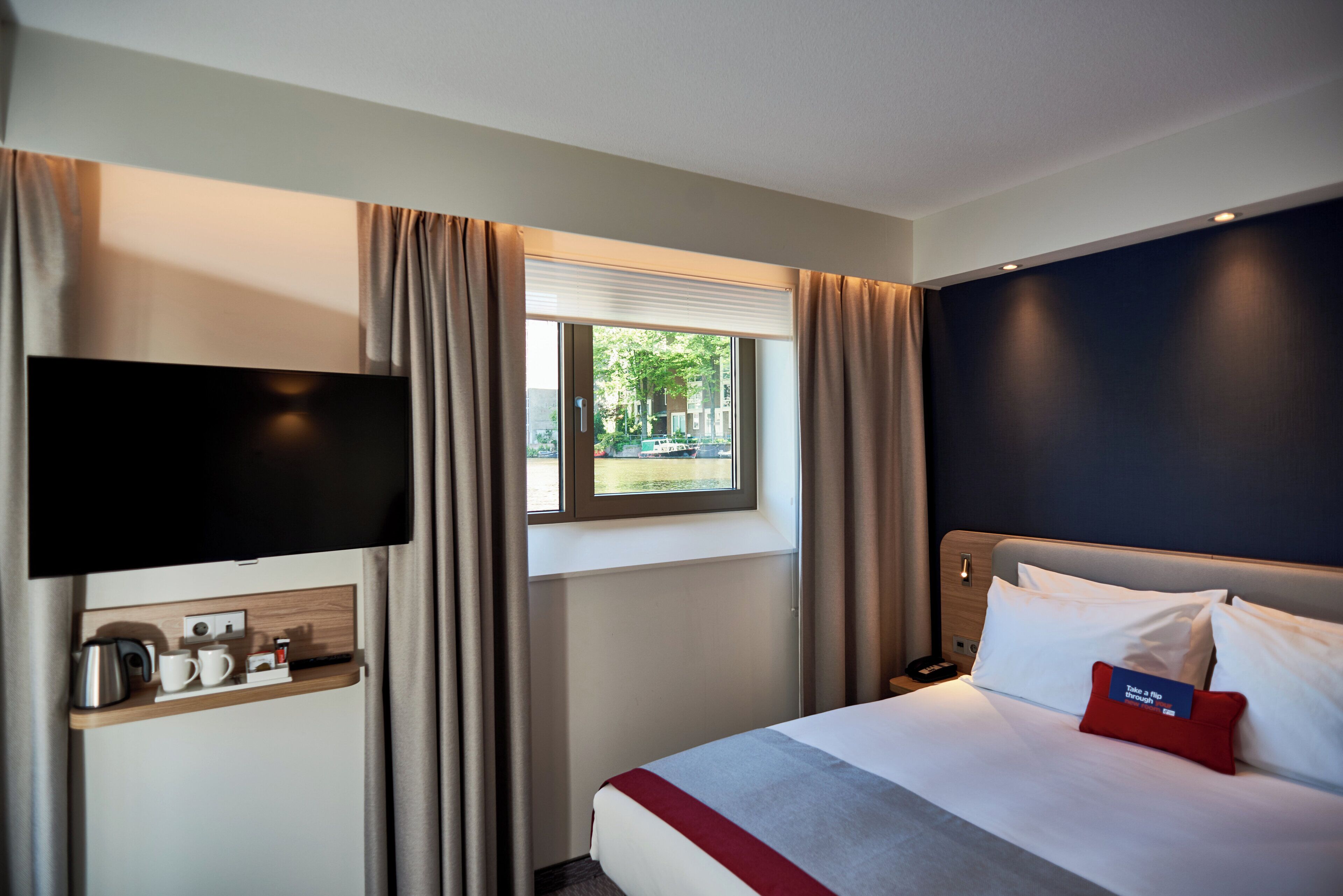 Ein Hotelzimmer mit Bett, Fernseher, Fenster mit Vorhängen und Nachttisch mit Wasserkocher und Tassen.