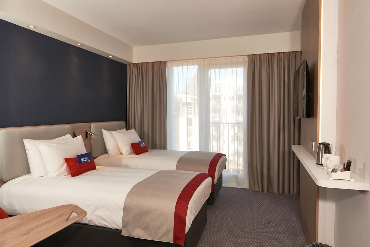 A hotel room with two beds, a flat-screen TV, a small desk, and a window with curtains.