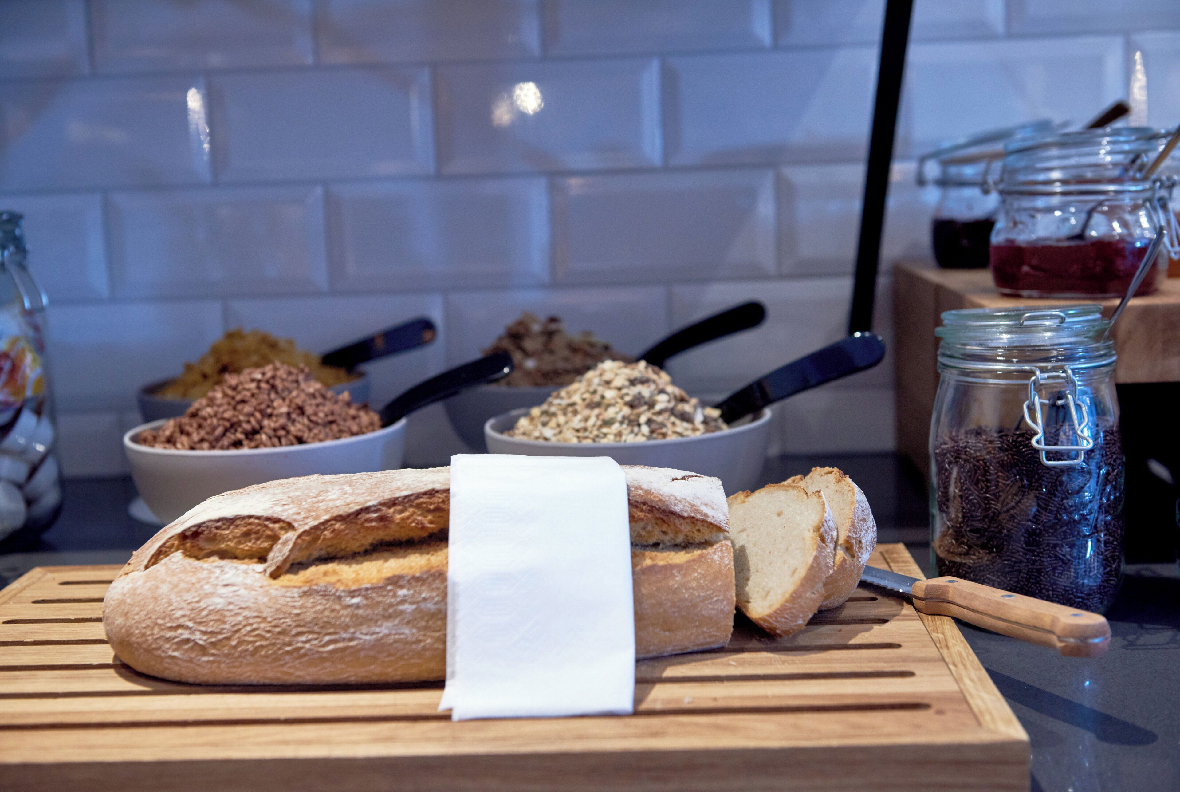 A loaf of bread on a cutting board with a knife, surrounded by bowls of grains and a jar of berries.