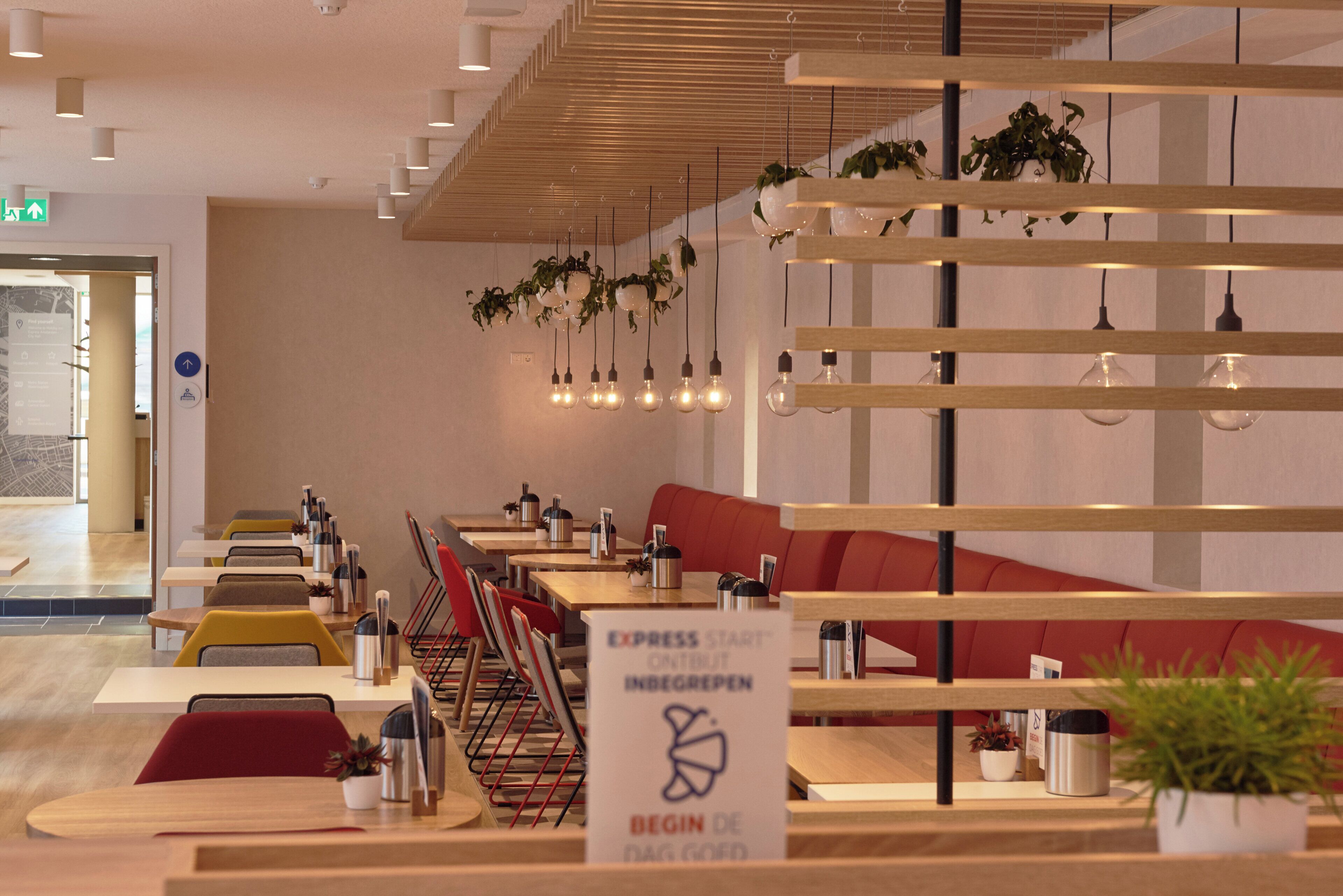Een modern restaurantinterieur met houten tafels, rode zitplaatsen, hangplanten en hanglampen.